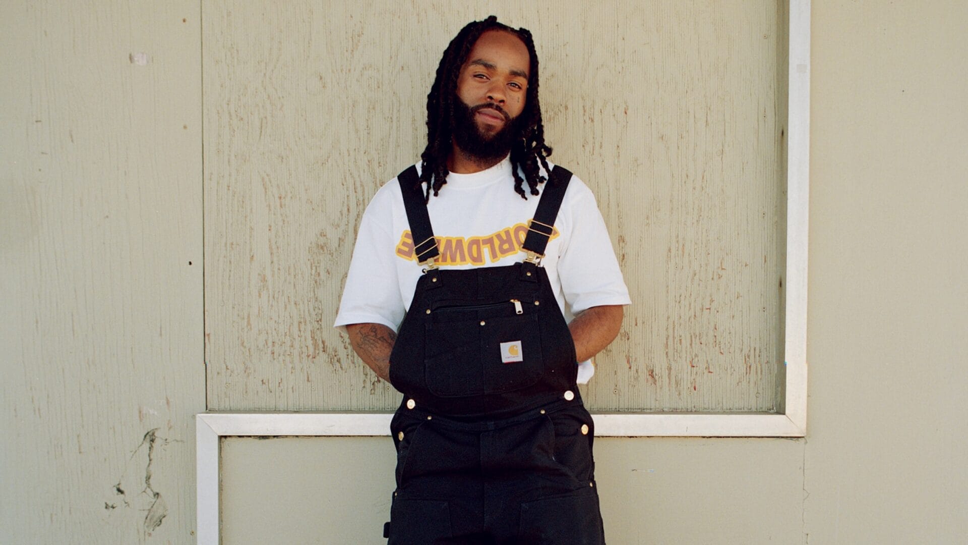 Fly Anakin standing in front of a pale cream colored wooden wall. He's wearing blue Carhartt overalls and a white t-shirt with "WORLDWIDE" written in mirror reverse on the chest.