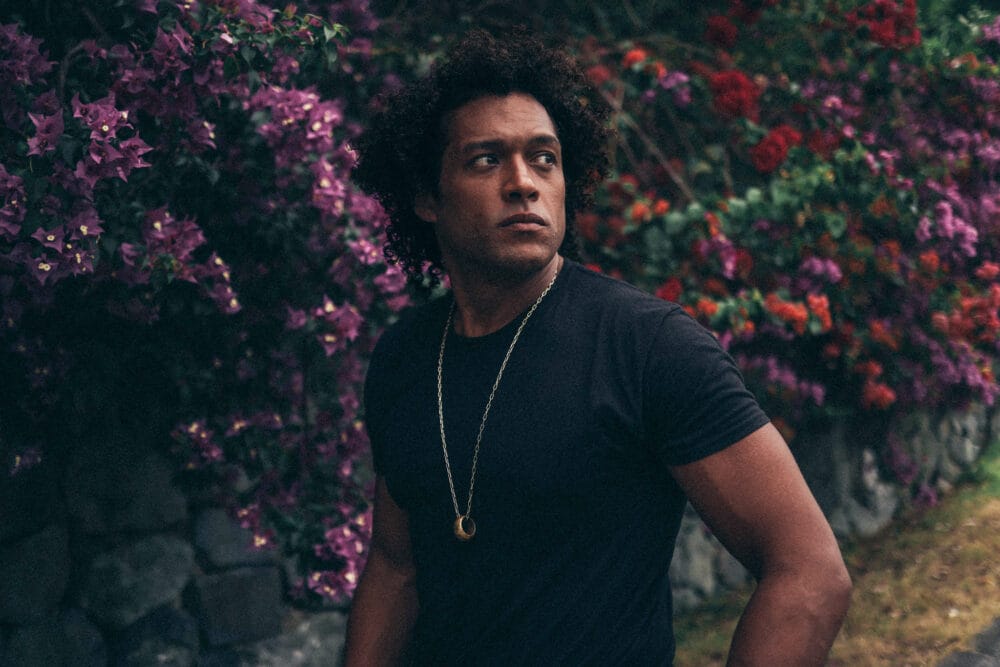 A dark skin man in a tight black tshirt and necklace with a ring on it, standing in front of a stone wall overgrown by purple and red flowers.