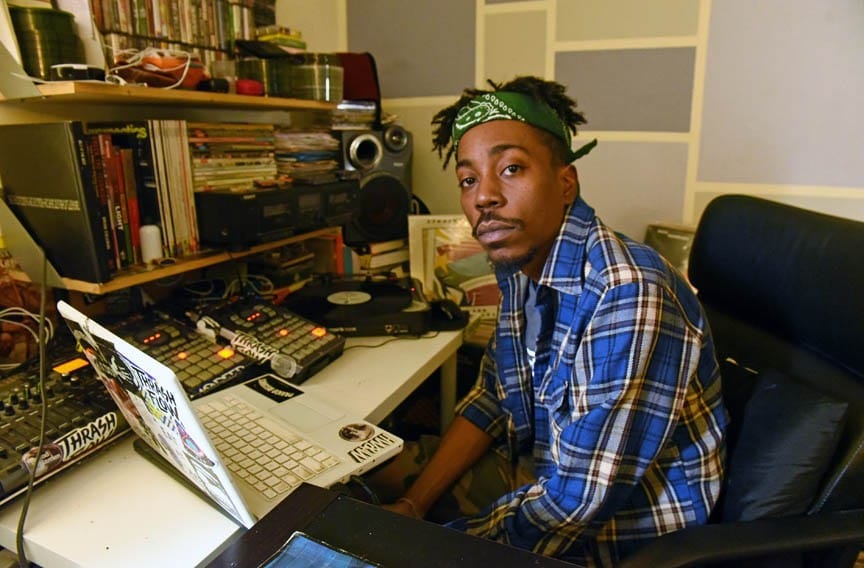 Obhliv sitting at his desk with several beat machines and a laptop. He's wearing a blue checkered flannel and a green bandana holding up his dreds.