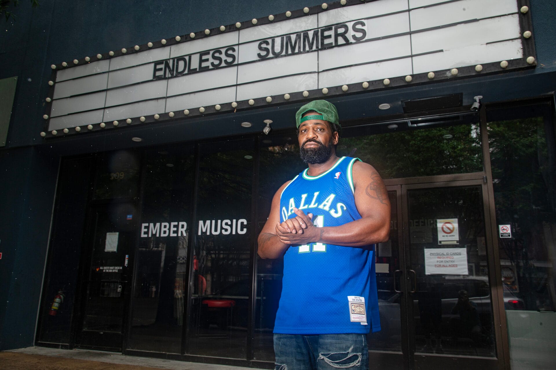 Nickelus F standing in front of a storefront, wearing a green snapback cap backwards and a Dallas Mavericks jersey.
