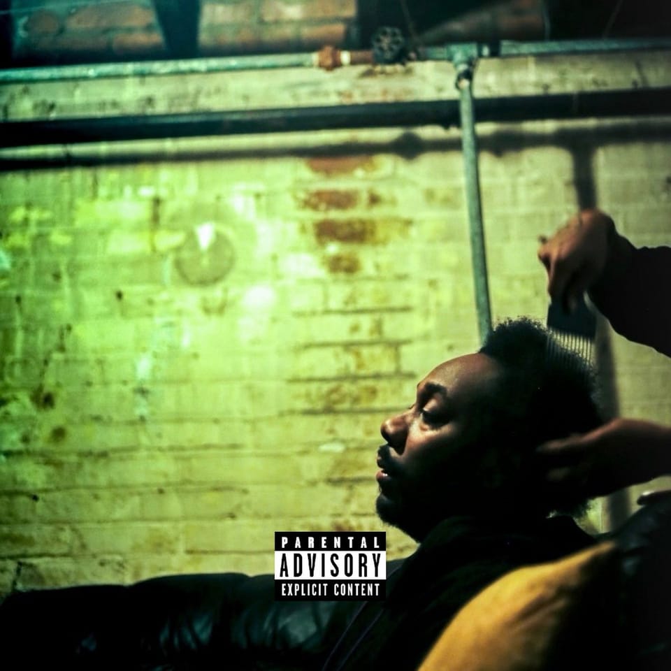 Kipp Stone sitting on a couch, having their afro picked out with a comb. They're in front of a brick wall in a basement, lit harshly. A 'parental advisory' logo is at the bottom of the frame.