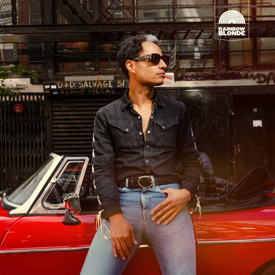 José James leaning languidly against a red covertible car parked in front of a city sidewalk backdrop. He's wearing wearing a black demin shirt, blue jeans, and sunglasses.