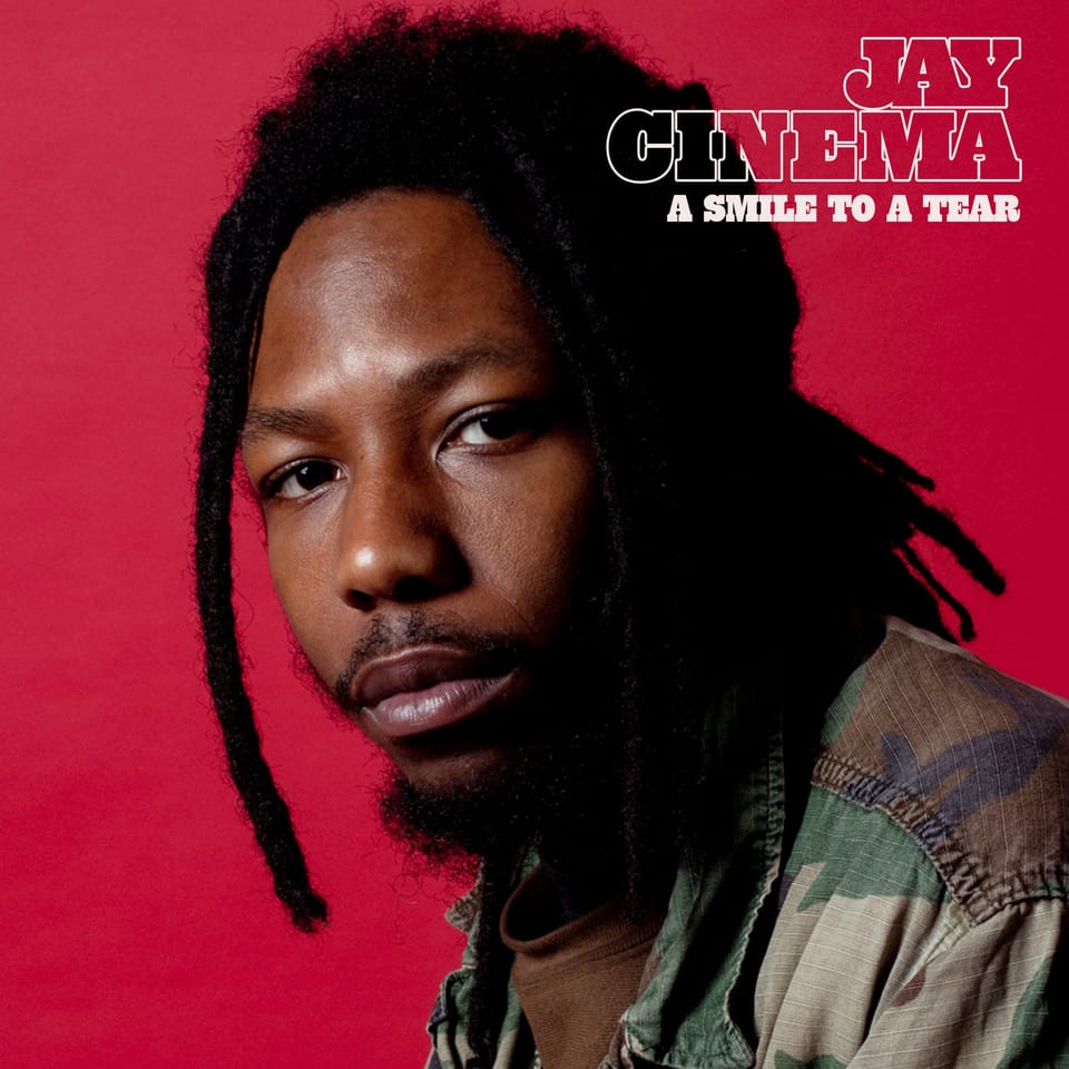 The cover for Jay Cinema's "A Smile To A Tear". Profile picture of a black man with dreds and a gotee looking toward the camera, in front of a plain red background.