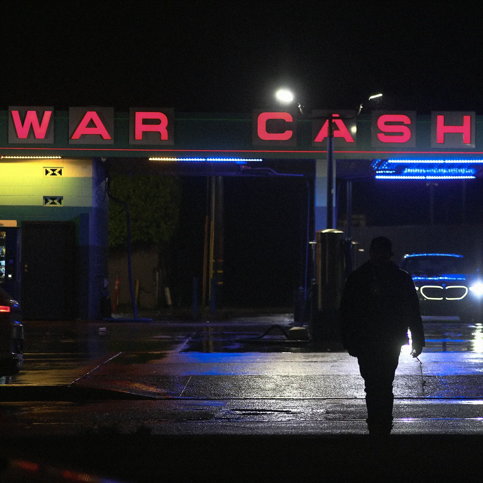 A car wash at night, its bright signage spells out "WAR CASH" in red lights. A silhouetted figure walks away from it toward the camera, a cigarette in hand.